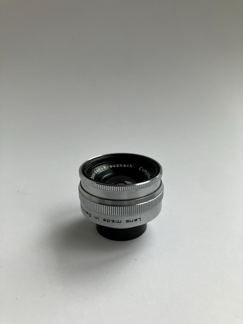 Puede incluir: Un objetivo de c&aacute;mara plateado y negro con el texto "Schneider-Kreuznach" y "Lens made in Germany" grabado en el lateral. El objetivo tiene un dise&ntilde;o estriado y un interior negro.