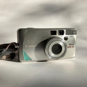 May include: Silver Canon Prima Super 155 camera with a zoom lens and attached strap. The camera has the Canon logo and a green accent. The camera is on a white background.