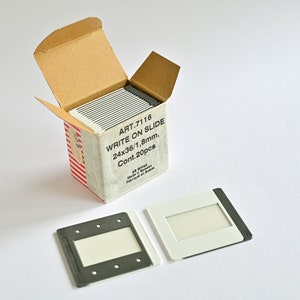 May include: A box of 20 blank 24x36 mm slides for writing on. The box is open and two slides are sitting next to it. One slide is white with a black border and the other is black with a white border.