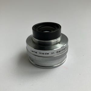 Puede incluir: Una lente de c&aacute;mara vintage con una parte superior negra y un cuerpo plateado. La lente est&aacute; marcada como "Lens made in Germany". La lente tiene un dise&ntilde;o estriado alrededor de la base.