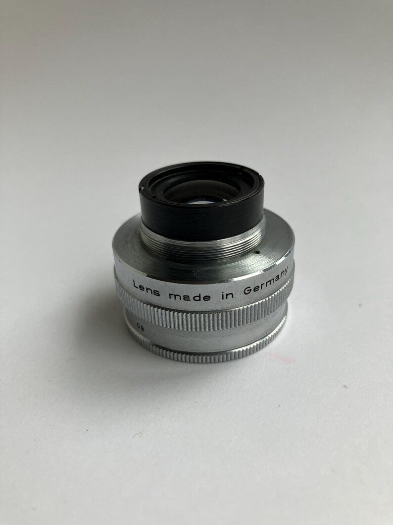 Puede incluir: Una lente de c&aacute;mara vintage con una parte superior negra y un cuerpo plateado. La lente est&aacute; marcada como "Lens made in Germany". La lente tiene un dise&ntilde;o estriado alrededor de la base.