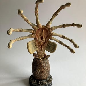 Puede incluir: Una escultura detallada de un facehugger Alien emergiendo de un huevo. El facehugger es de color beige con múltiples apéndices en forma de dedos. El huevo es marrón y texturizado, descansando sobre una base negra con la palabra "ALIEN" en blanco.