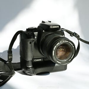 Könnte beinhalten: Eine schwarze Canon EOS Rebel 3000 Kamera mit einem Canon Zoom Lens EF 35-100mm 1:3.5-4.5 Objektiv. Die Kamera hat einen schwarzen Gurt.