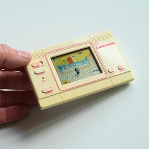 May include: A vintage handheld electronic game, cream-colored with pink accents, featuring a Cake Shop theme. The screen displays a pixelated game scene. Buttons include sound, select, and order. The game is held in a hand.