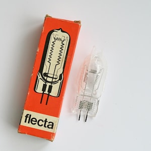 Puede incluir: Una caja de bombilla Flecta naranja y blanca con una bombilla transparente al lado. La caja tiene una ilustración en blanco y negro de la bombilla. La bombilla es transparente con dos clavijas negras.