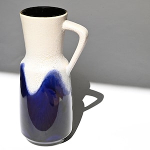 Puede incluir: Una jarra alta de cerámica con asa. La parte superior es blanquecina con una superficie texturizada, que pasa a un esmalte azul oscuro en la base. El interior es negro.