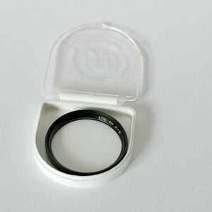 Pode incluir: Um filtro de lente de câmera preto e branco em uma caixa de plástico branca. O filtro é circular com uma borda preta e um centro transparente. A caixa está aberta, revelando o filtro dentro.