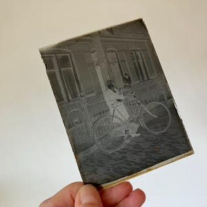 Może przedstawiać: Zdjecie w stylu vintage, czarno-białe, przedstawiające osobę jadącą na rowerze po brukowanej ulicy. Obraz przedstawia budynek z oknami i okiennicami w tle. Zdjęcie jest trzymane przez dłoń.