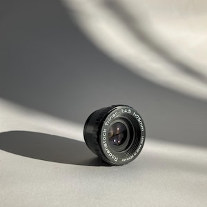 May include: A black Rodenstock Trinar 1:4.5 f=75mm camera lens. The lens is made in Germany and has white text on the black body. The lens is positioned on a white surface with a shadow cast to the left.