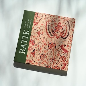 Könnte beinhalten: Ein Buch mit dem Titel "Batik" mit grünem Buchrücken und gemustertem Einband. Der Einband zeigt einen beigefarbenen Hintergrund mit roten und schwarzen Blumen- und Vogelmotiven. Das Buch liegt auf einer weißen Oberfläche.