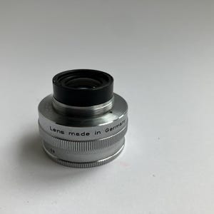 Puede incluir: Un objetivo de c&aacute;mara plateado con la parte superior negra. El objetivo est&aacute; marcado con "Lens made in Germany". El objetivo est&aacute; sobre una superficie blanca.
