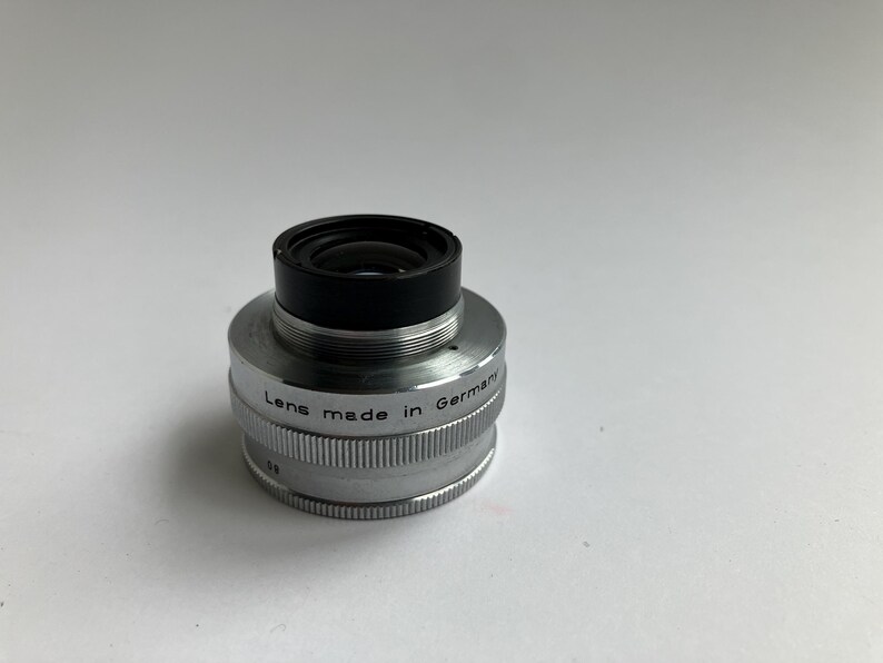 Puede incluir: Un objetivo de c&aacute;mara plateado con la parte superior negra. El objetivo est&aacute; marcado con "Lens made in Germany". El objetivo est&aacute; sobre una superficie blanca.