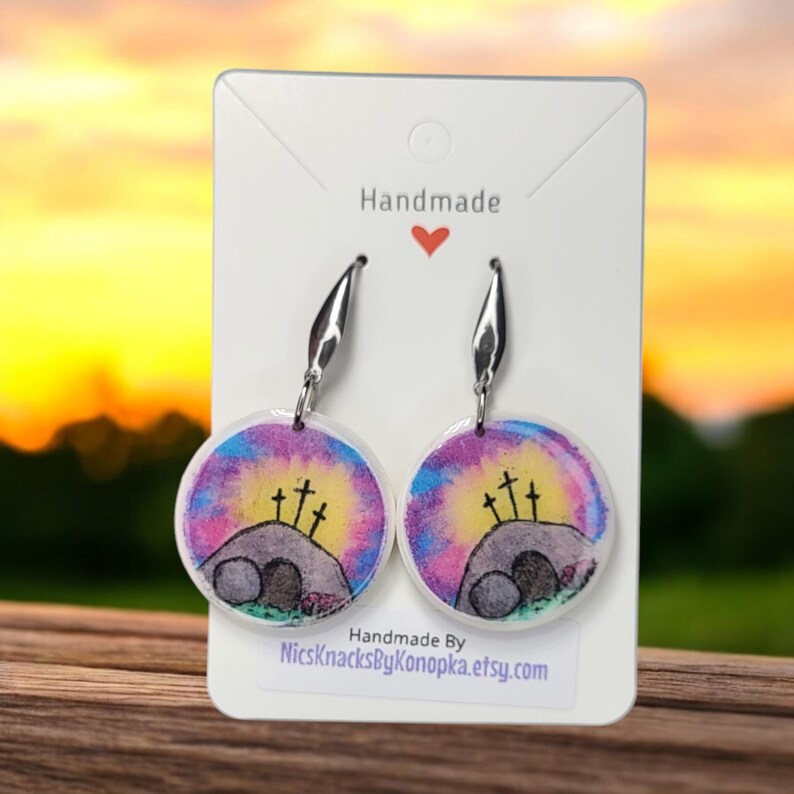 May include: Handmade dangle earrings with a watercolour design of a hill with three crosses and an empty tomb. The earrings have silver-tone hooks and are displayed on a white card with the word "Handmade".