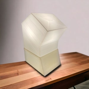 May include: A white, geometric lamp with a unique, angular design. The lamp is made of a smooth, textured material and has a base that is slightly wider than the top. The lamp is sitting on a wooden table.