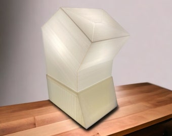Op de afbeelding: Een witte, geometrische lamp met een uniek, hoekig ontwerp. De lamp is gemaakt van een glad, getextureerd materiaal en heeft een voet die iets breder is dan de bovenkant. De lamp staat op een houten tafel.