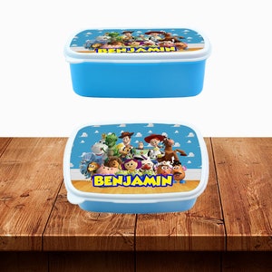 Op de afbeelding: Blauwe plastic lunchbox met een kleurrijk beeld van personages uit de Toy Story-filmfranchise. De lunchbox heeft een wit deksel met de naam "BENJAMIN" erop gedrukt.