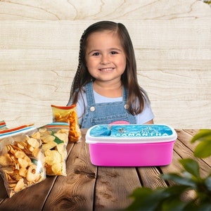 May include: A pink lunchbox with a blue and white lid featuring a Frozen character and the name "Samantha". The lunchbox is on a wooden table with bags of chips.