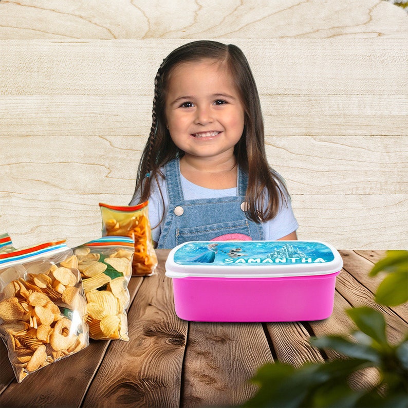 May include: A pink lunchbox with a blue and white lid featuring a Frozen character and the name "Samantha". The lunchbox is on a wooden table with bags of chips.