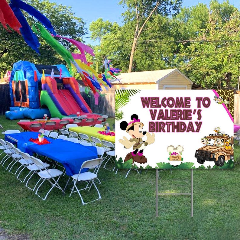 Peut inclure: Une pancarte d'accueil blanche avec de la v&eacute;g&eacute;tation verte et une Minnie Mouse de dessin anim&eacute; portant une tenue de safari. La pancarte indique "Bienvenue &agrave; l'anniversaire de Val&eacute;rie". Il y a une jeep de dessin anim&eacute; avec un tigre, un lion et un singe &agrave; l'int&eacute;rieur. La pancarte est sur un support m&eacute;tallique dans une cour avec un ch&acirc;teau gonflable et des tables avec des chaises.
