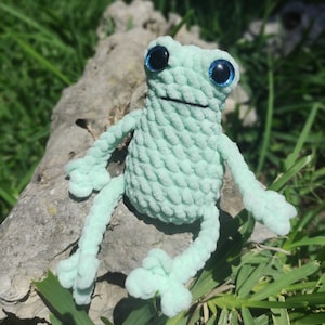 May include: A crocheted green frog with blue eyes sitting on a rock. The frog is made of soft yarn and has a textured surface.