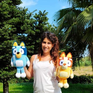 May include: Two handmade crochet plush toys, one blue and one yellow, resembling cartoon characters. The blue toy has a light blue body, dark blue head, and yellow accents. The yellow toy has a yellow body, orange head, and white accents. The toys are held by a person.