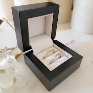 May include: Two delicate rings with small diamonds set in a black ring box. The ring on the left is gold and the ring on the right is silver.