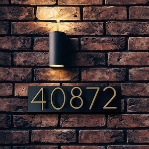 May include: A black rectangular sign with gold numbers "40872" mounted on a brick wall. A black light fixture with a light bulb is mounted above the sign.