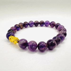 May include: A purple amethyst gemstone bracelet with a gold charm. The bracelet is made of round beads and has a clasp.