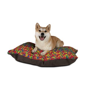 May include: A Shiba Inu dog rests on a colorful dog bed. The bed has a brown base and a top covered in a pattern of red, yellow, blue, and green shapes. The dog is tan and white with a happy expression.