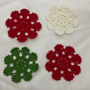 Puede incluir: Cuatro posavasos de copo de nieve hechos a crochet en rojo, blanco y verde. Los posavasos están hechos de hilo y tienen un diseño delicado y de encaje.