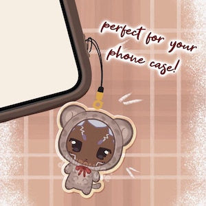 May include: A phone charm featuring a cartoon bear character in a hooded outfit with a red bow. The charm is attached to a phone case. Text reads "perfect for your phone case!" and "preorders open 11.15.25".