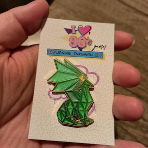 May include: A green geometric dragon enamel pin with gold trim, on a white card with a purple design. The card reads "I ❤️ 90's games" and "@jessie_inkswell".