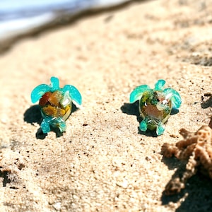 May include: Two teal blue resin turtle earrings with a variety of colorful sand and shells embedded inside. The earrings are on a sandy beach.