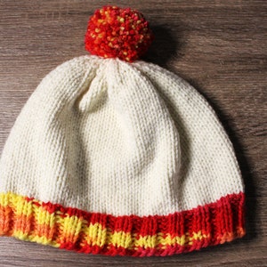 K&ouml;nnte beinhalten: Eine wei&szlig;e Strickm&uuml;tze mit einem roten und gelben Bommel oben. Die M&uuml;tze hat ein rotes, gelbes und oranges Band um den unteren Rand.