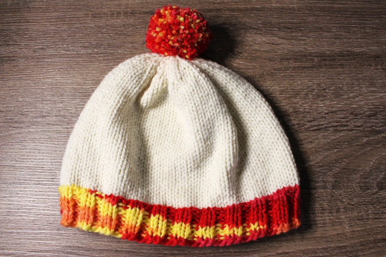 K&ouml;nnte beinhalten: Eine wei&szlig;e Strickm&uuml;tze mit einem roten und gelben Bommel oben. Die M&uuml;tze hat ein rotes, gelbes und oranges Band um den unteren Rand.