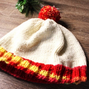 K&ouml;nnte beinhalten: Eine wei&szlig;e Strickm&uuml;tze mit einem rot-gelb-orangenen gestreiften Rand und einem roten Bommel oben.