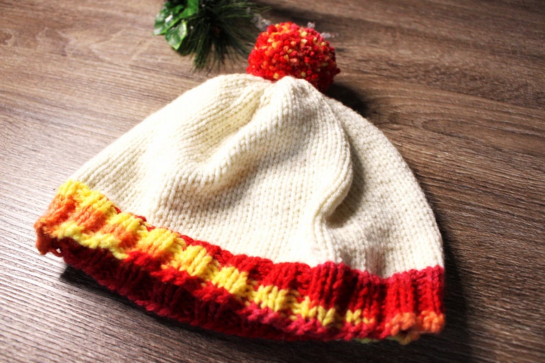 K&ouml;nnte beinhalten: Eine wei&szlig;e Strickm&uuml;tze mit einem rot-gelb-orangenen gestreiften Rand und einem roten Bommel oben.