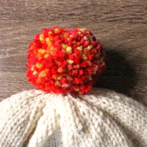 K&ouml;nnte beinhalten: Wei&szlig;e Strickm&uuml;tze mit einem roten, orangen und gelben Bommel oben.