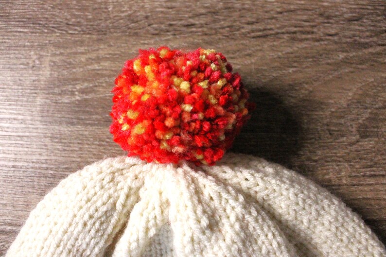 K&ouml;nnte beinhalten: Wei&szlig;e Strickm&uuml;tze mit einem roten, orangen und gelben Bommel oben.