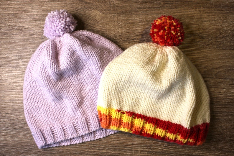 K&ouml;nnte beinhalten: Zwei gestrickte M&uuml;tzen mit Bommeln. Eine M&uuml;tze ist helllila und die andere ist cremefarben mit einem rot-orange-gelben Streifenband.