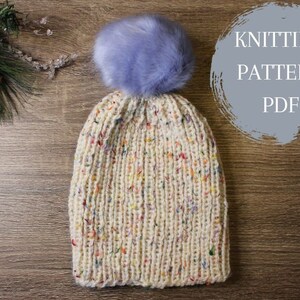 Pode incluir: Um chapéu branco de tricô com um padrão salpicado de cores e um pompom de pele sintética roxo claro. O chapéu está sobre uma superfície de madeira com o texto "Knitting Pattern PDF" em um círculo.