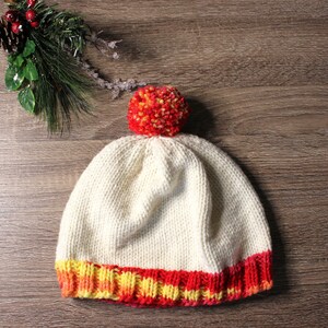 K&ouml;nnte beinhalten: Eine wei&szlig;e Strickm&uuml;tze mit einem roten und gelben Bommel und einem roten, gelben und orangenen gestreiften Rand.