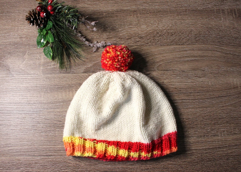 K&ouml;nnte beinhalten: Eine wei&szlig;e Strickm&uuml;tze mit einem roten und gelben Bommel und einem roten, gelben und orangenen gestreiften Rand.