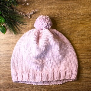 K&ouml;nnte beinhalten: Eine rosa gestrickte M&uuml;tze mit einem flauschigen Bommel oben. Die M&uuml;tze liegt auf einer Holzoberfl&auml;che.
