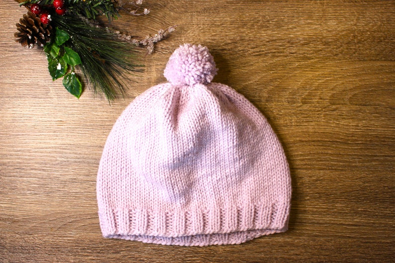 K&ouml;nnte beinhalten: Eine rosa gestrickte M&uuml;tze mit einem flauschigen Bommel oben. Die M&uuml;tze liegt auf einer Holzoberfl&auml;che.