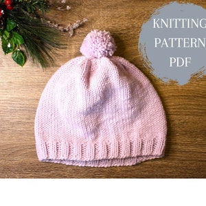 Op de afbeelding: Een roze gebreide muts met een witte pompon bovenop. De muts ligt op een houten oppervlak. De tekst "Knitting Pattern PDF" staat op een grijze cirkel in de rechterbovenhoek van de afbeelding.