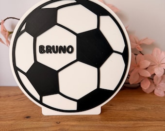 Könnte beinhalten: Eine kreisförmige Dekoplatte mit einem schwarz-weißen Fußballmuster. Der Name BRUNO ist in schwarzen Großbuchstaben in der Mitte des Balls eingeprägt. Das Objekt ist auf einem flachen weißen Sockel für eine aufrechte Präsentation montiert.