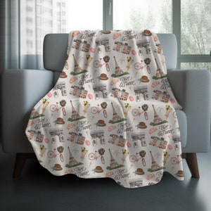 May include: A cream-colored throw blanket draped over a chair, featuring a repeating pattern of Parisian landmarks. The design includes the Eiffel Tower, Arc de Triomphe, bicycles, hats, and the phrase "Vive la France" in script.