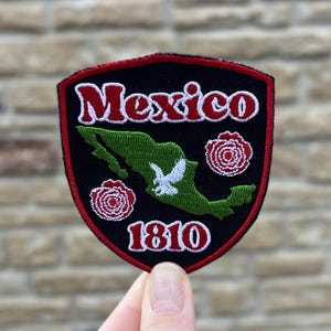 May include: A black and red embroidered patch with the word "Mexico" in red script. The patch features a green map of Mexico, white bird, and the year "1810". Two red roses are on the patch.