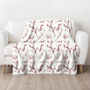 May include: A white throw blanket featuring a repeating pattern of dark brown branches with clusters of small red berries. The design is spread across the fabric, showcasing a botanical illustration style with thin stems and round red fruit elements.